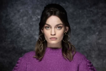 A Beauty Shot Of The Famous Actress, Emma Mackey, Sporting A Bouffant Hairstyle, Gold Hoop Earrings, And A Purple Sweater.