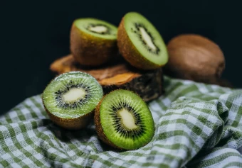 Hd Still Life Photography A Set Of Kiwis Cut In Half Laid On Top A Small Tree Trunk And A Green Checkered Tablecloth.
