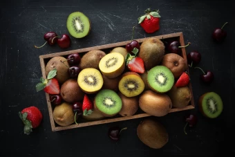 Top View Shot Of A Fruit Box Comprising Of Whole And Halved Yellow And Green Kiwis, Cherries, And Strawberries Showcased Before A Dark Scratched Surface