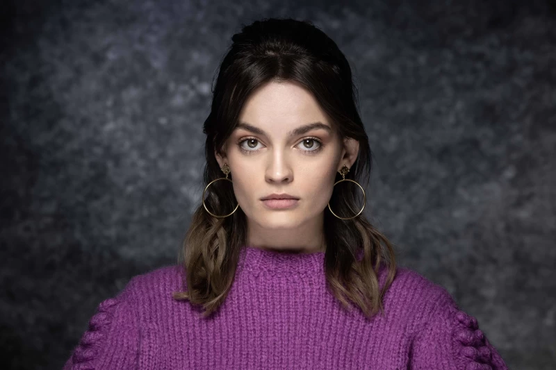 A Beauty Shot Of The Famous Actress, Emma Mackey, Sporting A Bouffant Hairstyle, Gold Hoop Earrings, And A Purple Sweater.