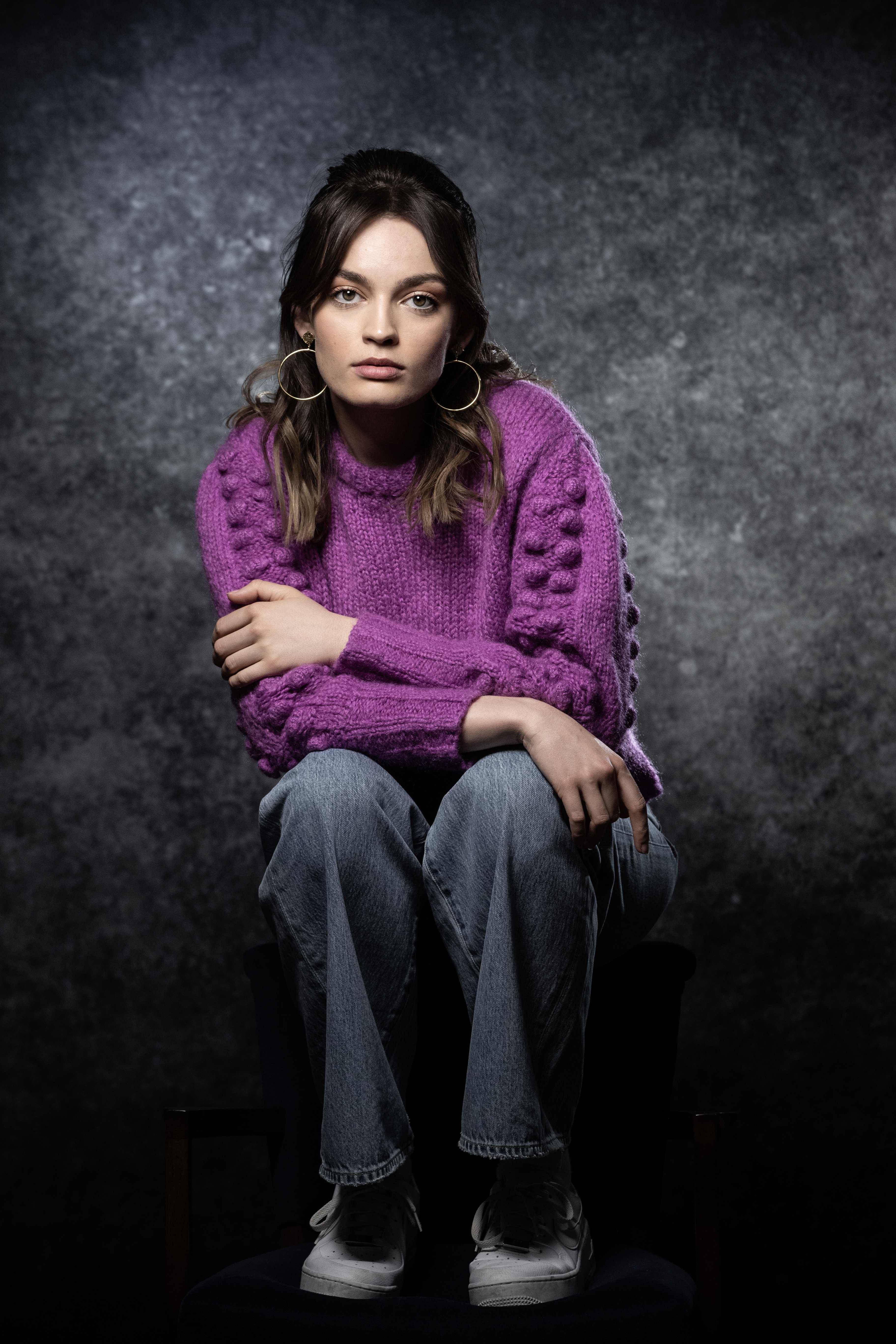 A Studio Portrait Of The Actress, Emma Mackey, Wearing A Distinct Purple Sweater While Sitting On A Chair