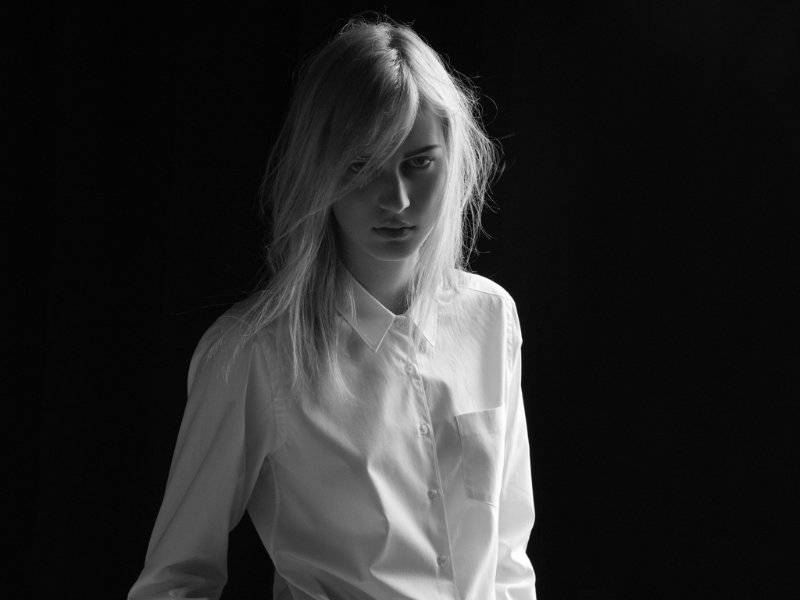 A Melancholic Shot Of Julia Nobis In Black And White As She Posed In Front Of A Black Backdrop While Wearing A Long White Sleeved Shirt