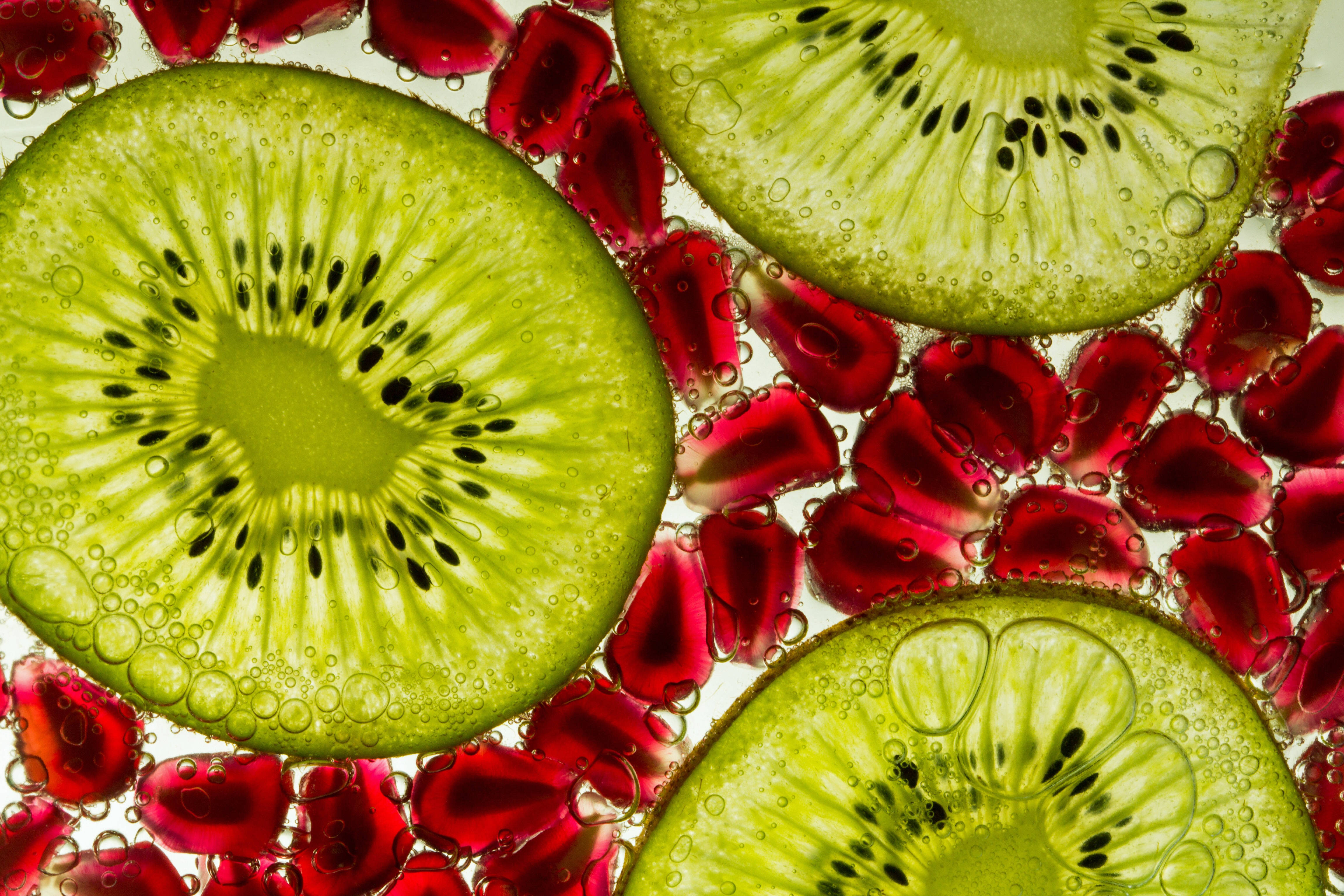 A Fascinating Macro Photography Image Of Red Translucent Pomegranate Seeds In The Spaces Between Three Slices Of The Kiwi Fruit Backlit From A White Surface